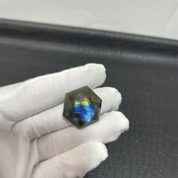 Cristal hexagonal en labradorite naturelle aux reflets arc-en-ciel vibrant, tenu par une main gantée sur un fond noir.