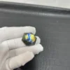 Cristal hexagonal en labradorite naturelle avec des reflets bleu et jaune, tenu par une main gantée blanche sur un fond texturé noir.