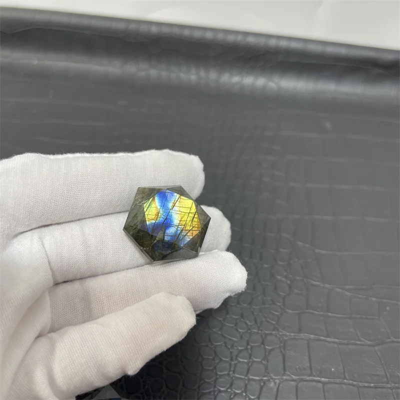 cristal-hexagonal-labradorite-naturelle-reflets-bleu-jaune-15 Cristal hexagonal en labradorite naturelle avec des reflets bleu et jaune, tenu par une main gantée blanche sur un fond texturé noir.