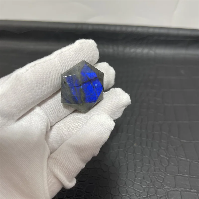 cristal-hexagonal-labradorite-naturelle-reflets-bleus-11 Cristal hexagonal en labradorite naturelle avec reflets bleus éclatants, tenu par une main gantée sur fond texturé noir.