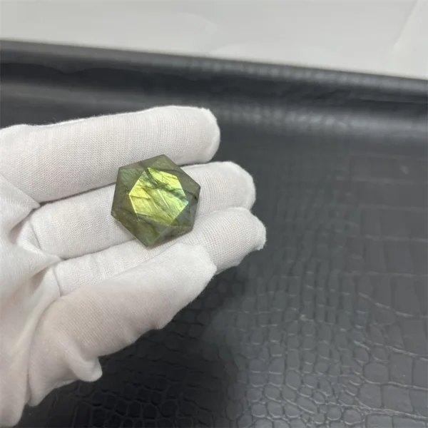 Cristal hexagonal en labradorite naturelle aux reflets jaunes, tenu par une main gantée blanche sur un fond noir texturé.