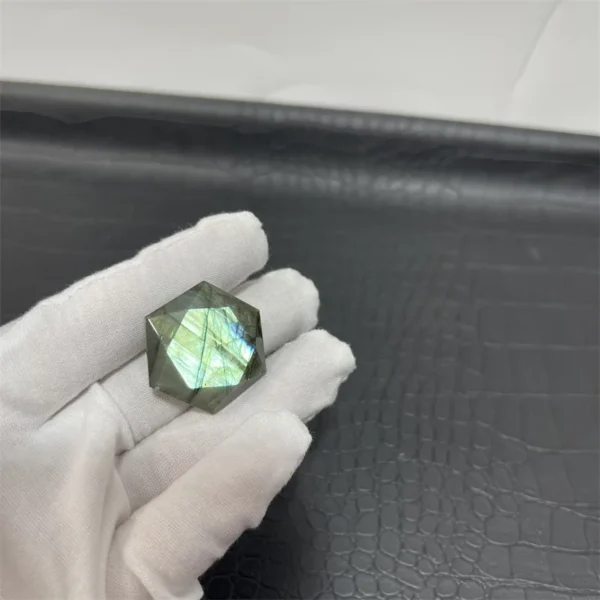 Cristal hexagonal en labradorite naturelle avec des reflets verts lumineux, tenu par une main gantée sur fond texturé noir.