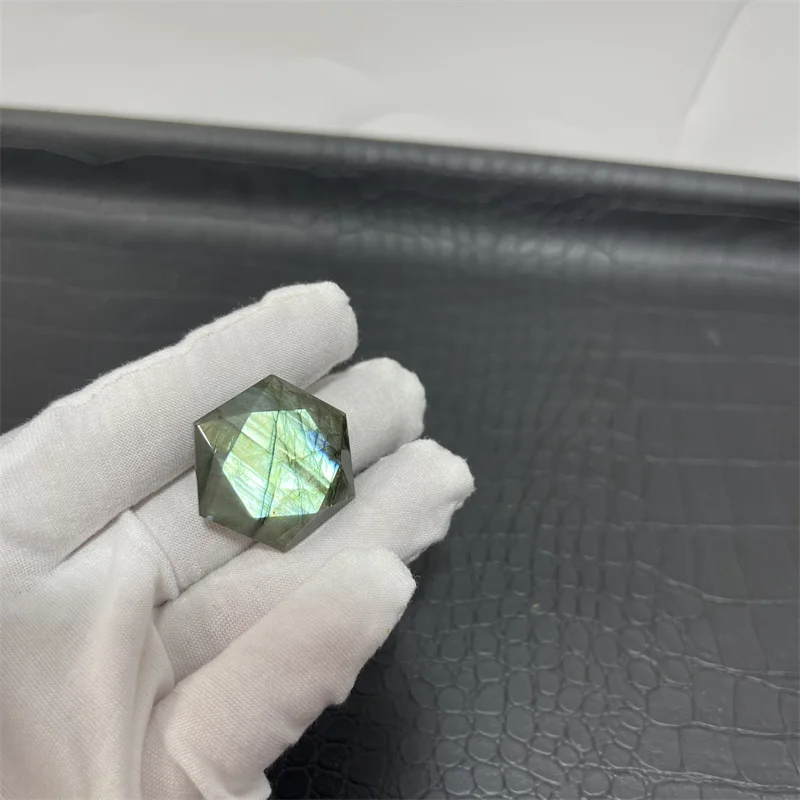 cristal-hexagonal-labradorite-naturelle-reflets-verts-12 Cristal hexagonal en labradorite naturelle avec des reflets verts lumineux, tenu par une main gantée sur fond texturé noir.
