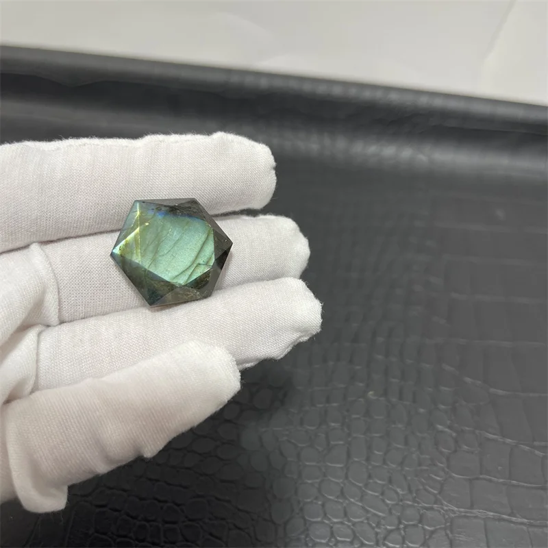 cristal-hexagonal-labradorite-naturelle-reflets-verts-13 Cristal en labradorite hexagonal avec des reflets verts, tenu par une main gantée blanche sur un fond noir texturé.