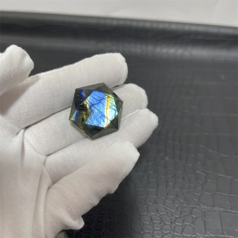 cristal-labradorite-hexagonal-reflets-bleus-17 Cristal de labradorite hexagonal avec des reflets bleus irisés, tenu par une main gantée sur un fond texturé.